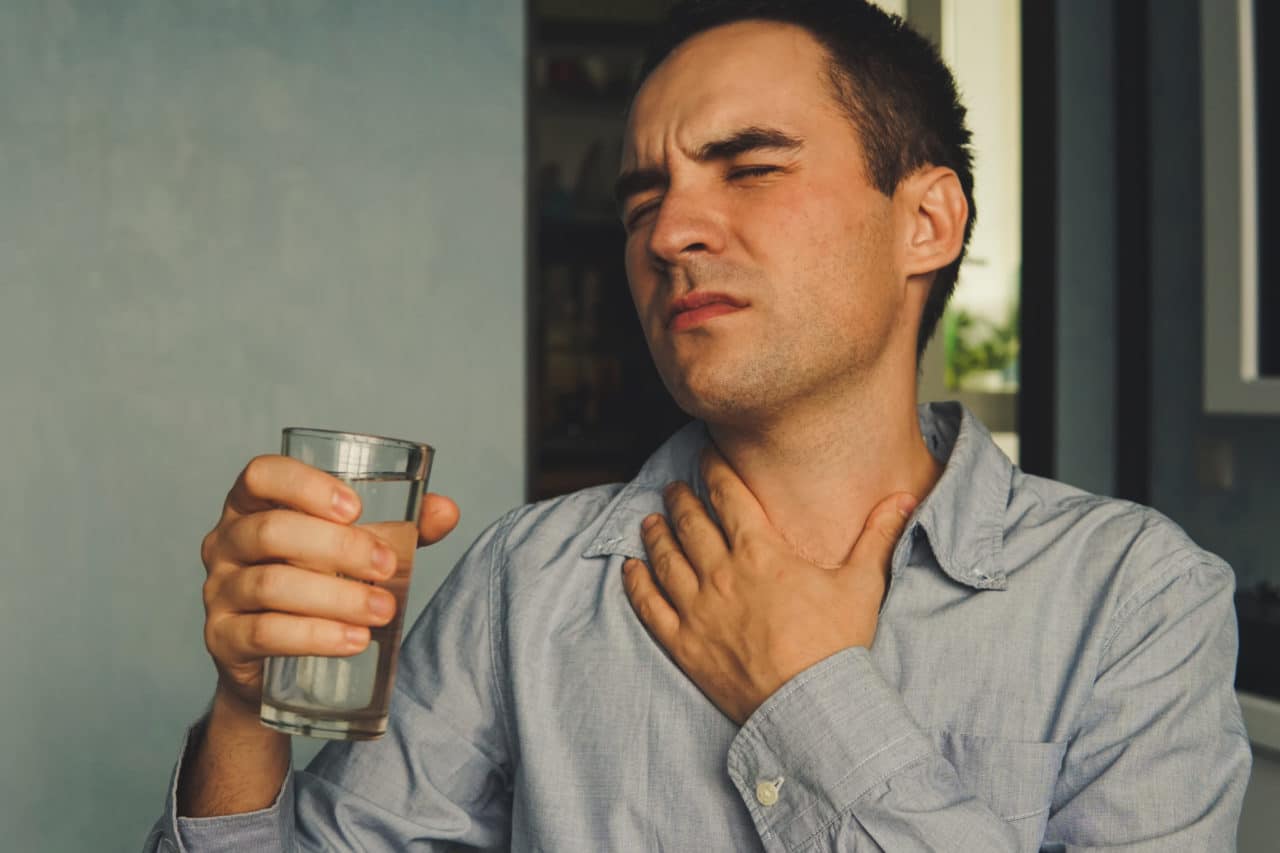 A person holding their throat and wincing in pain while holding a glass of water