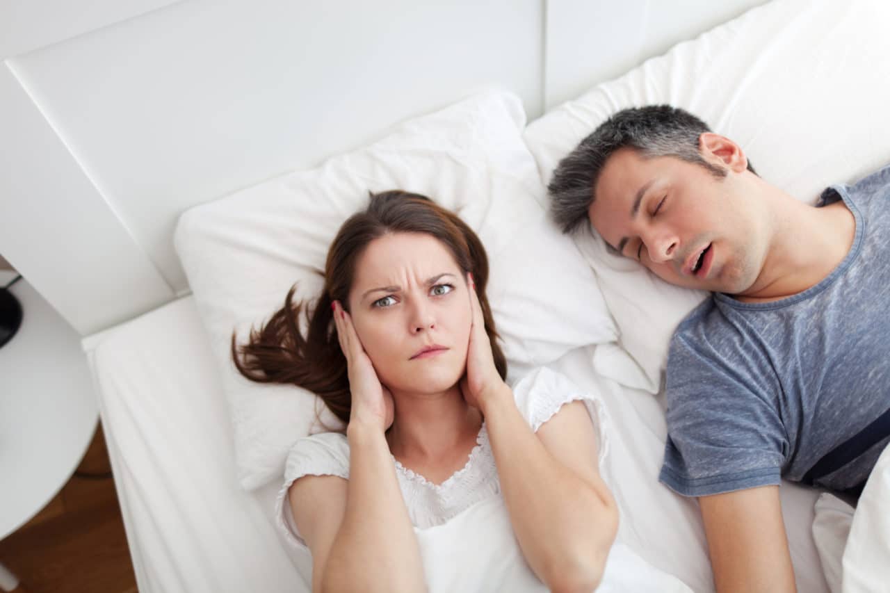 A person looking annoyed and holding their hands over their ears while the other person in the bed sleeps, presumably snoring