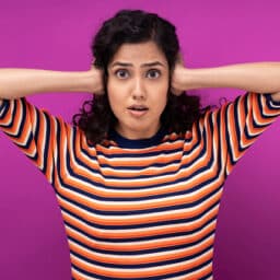 Woman standing against a pink background covering her ears.