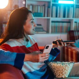 Young woman relaxing at home, eating popcorn and using phone while watching TV