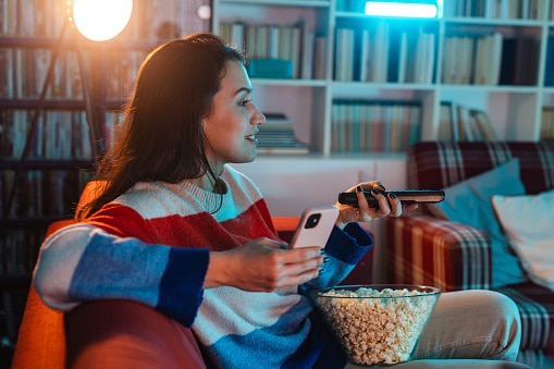 Young woman relaxing at home, eating popcorn and using phone while watching TV.