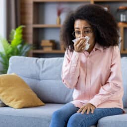 Woman sitting on her couch, blowing her nose into a tissue.