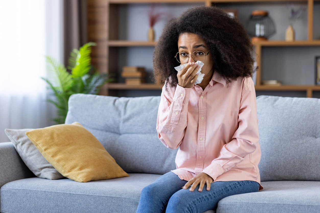 Woman sitting on her couch, blowing her nose into a tissue.