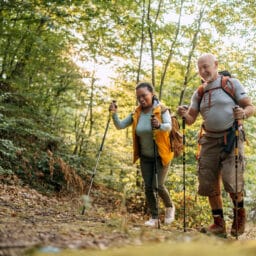 A smiling older couple hikes along a forest trail with trekking poles and backpacks, enjoying nature and a healthy outdoor activity together on a sunny day.