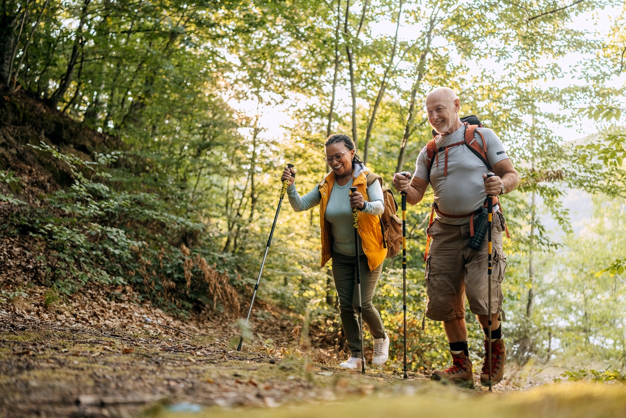 A smiling older couple hikes along a forest trail with trekking poles and backpacks, enjoying nature and a healthy outdoor activity together on a sunny day.